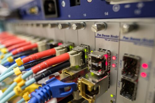 Nahaufnahme eines Netzwerkracks mit Glasfaser- und Ethernet-Anschlüssen. Mehrere farbige Kabel, darunter rote, blaue und gelbe, sind in die Ports eingesteckt. Die Netzwerk-Ports sind mit LEDs ausgestattet, von denen einige rot und grün leuchten, was auf die Aktivität und den Status der Verbindungen hinweist.