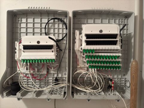 Dieses Bild zeigt zwei nebeneinander installierte Glasfaserverteilerboxen. Jede Box enthält mehrere Glasfaseranschlüsse und -verteiler, die jeweils mit grünen SC/APC-Adapterports versehen sind, um die optischen Fasern miteinander zu verbinden. Die Verkabelung ist sorgfältig organisiert, wobei die roten und weißen Glasfaserkabel an den Ports angebracht sind. Auf der linken Seite der Box befinden sich schwarze Kabeltüllen, die ungenutzte Anschlüsse abdecken oder für zukünftige Erweiterungen vorgesehen sein könnten.